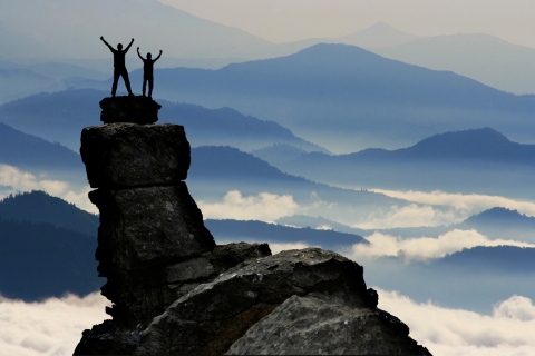 Zyskasz pewność siebie w mówieniu Zdjęcie przedstawia dwie osoby stojące na szczycie wysokiej skały, triumfalnie unoszące ręce w górę. Za nimi rozciąga się malowniczy krajobraz gór, otoczonych mgłą i chmurami. Obraz symbolizuje pokonywanie własnych ograniczeń, osiąganie celów i zdobywanie pewności siebie.