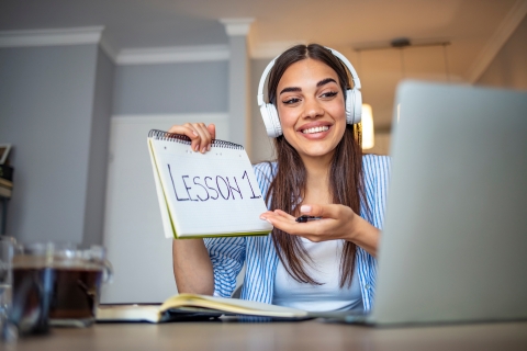 Zdjęcie przedstawia uśmiechniętą młodą kobietę uczestniczącą w lekcji online. Ma na sobie słuchawki i pokazuje notatnik z napisem „Lesson 1”, co sugeruje, że właśnie rozpoczyna naukę. Obraz symbolizuje, że kursy są przeznaczone dla osób, które chcą uczyć się w wygodny, elastyczny sposób – zarówno dla początkujących, jak i tych, którzy pragną rozwijać swoje umiejętności niezależnie od miejsca i czasu.