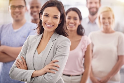 Na zdjęciu widać uśmiechniętą kobietę w eleganckim, jasnoszarym garniturze, stojącą na pierwszym planie z założonymi rękami. Za nią znajduje się grupa ludzi, którzy również się uśmiechają i wyglądają na zadowolonych. Ich różnorodność sugeruje zespół pracujący w przyjaznej i profesjonalnej atmosferze. Obrazek symbolizuje gotowość do dostosowania kursu do specyficznych potrzeb firmy oraz podkreśla znaczenie skutecznego i dobrze dobranego szkolenia dla zespołu.